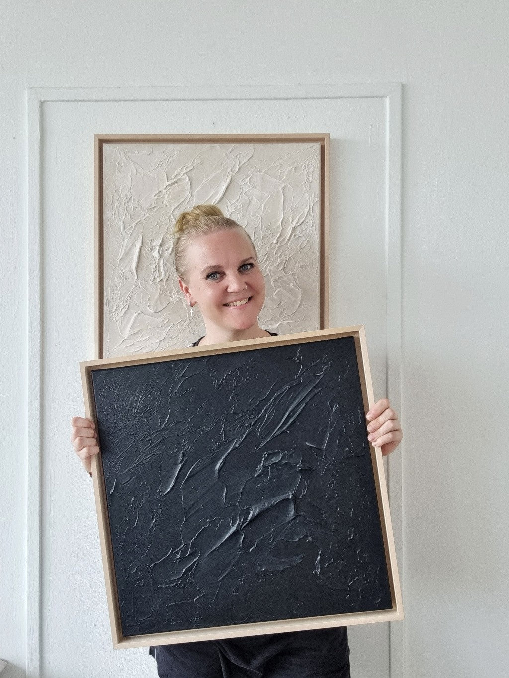 Female artist in the studio holding black monochrome textured painting framed with wooden floating frames.